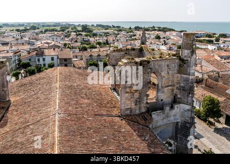 Le rovine del transetto medievale della chiesa di Saint-Martin si ergono accanto al tetto della chiesa in Place Eudes d'Aquitaine a Saint-Martin-de-Ré sull'isola di Ré (Île de Ré), Charente-Maritime, Nouvelle-Aquitaine, Francia. Dietro le mura con archi di finestre gotici e una torretta appuntita, i tetti dalle tegole rosse della città costiera si estendono fino al mare. La chiesa fortificata gotica fu costruita nei secoli XIV e XV. Nel 1696, una flotta anglo-olandese la devastò con il suo bombardamento. Durante la ricostruzione nel XVIII secolo, fu costruito un nuovo campanile, e le rovine del transetto con il suo camminamento Foto Stock