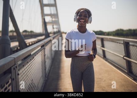 Donna afro-americana felice e in forma con abbigliamento sportivo, jogging e ascolto di musica attraverso le cuffie sul ponte urbano della città. Foto Stock