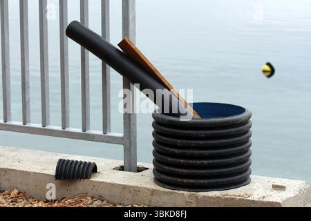 Il tubo corrugato nero e il pannello di legno poggiano in un grande anello di plastica su un bordo di cemento accanto a una recinzione metallica, con una boa galleggiante in acqua calma Foto Stock