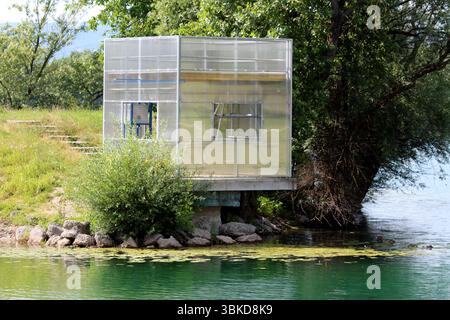 Piccolo e moderno rifugio con pareti in policarbonato traslucido e finestre aperte si erge sull'acqua, circondato da vegetazione lussureggiante e rocce, che si riflettono nel Foto Stock