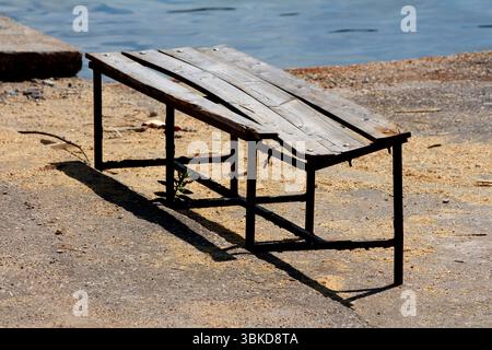 Vecchia panchina in legno con telaio in metallo piegato e tavole irregolari si trova su una superficie di cemento vicino al bordo dell'acqua, gettando un'ombra al sole e mostrando S. Foto Stock