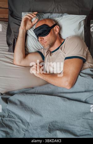 Ritratto di uomo calvo di mezza età con barba corta che indossa una maschera dormiente nera comodamente a letto durante la mattina presto promuovendo un sano riposo Foto Stock