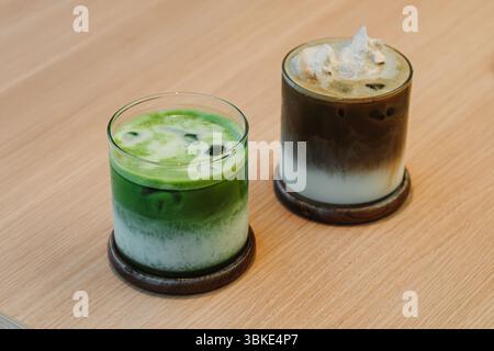Due bevande a strati ghiacciati, latte matcha e hojicha latte, serviti in bicchieri di vetro trasparente su sottobicchieri di legno, posti su un tavolo di legno chiaro Foto Stock