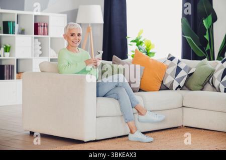 Luminoso salotto con una donna anziana che si rilassa su un comodo divano che regge uno smartphone e mostra un atteggiamento allegro Foto Stock