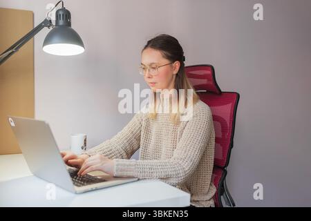 Donna che scrive su un computer portatile sotto la lampada da scrivania nell'ufficio domestico con sedia in rete rossa. Lavoratrice libera seduta alla scrivania bianca, indossando un maglione beige Foto Stock