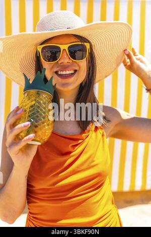 Donna felice che indossa un cappello di paglia e occhiali da sole con un cocktail a forma di ananas davanti a uno sfondo a righe bianche e gialle Foto Stock