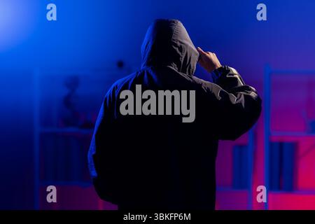 Vista posteriore di un uomo in una felpa con cappuccio nera in piedi in una stanza buia illuminata al neon, che tira giù la felpa con cappuccio per coprirgli il viso, evocando temi di sicurezza informatica, Foto Stock