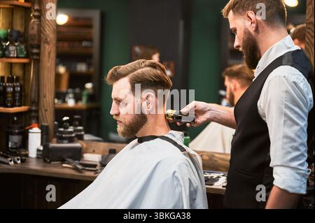 Bell'uomo in visita al parrucchiere in barbiere. Parrucchiere professionale che utilizza un rasoio elettrico per radere la pelle con cura. Vista laterale del cliente tagliato di capelli sullo sfondo dello specchio grande. Foto Stock