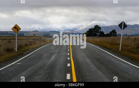 Segui le indicazioni per Winding Road. Montagne innevate in lontananza. Centro di Otago. Isola del Sud. Nuova Zelanda. Foto Stock