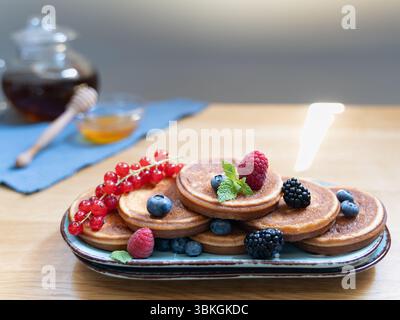 Pancake su un piatto blu ricoperto di frutti di bosco e banana, con miele e teiera di vetro sullo sfondo, la plafoniera mette in risalto la scena Foto Stock