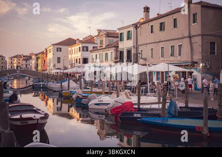Chioggia, italia 21 giugno 2025, persone che cenano in ristoranti all'aperto lungo un canale di chioggia, italia, con barche ormeggiate nelle vicinanze durante un bel sole Foto Stock