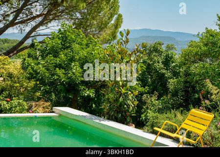 Una piscina con acqua verde smeraldo in Provenza (Francia) immersa in Un lussureggiante paesaggio mediterraneo. Una sedia da giardino gialla si trova al bordo della piscina. Foto Stock