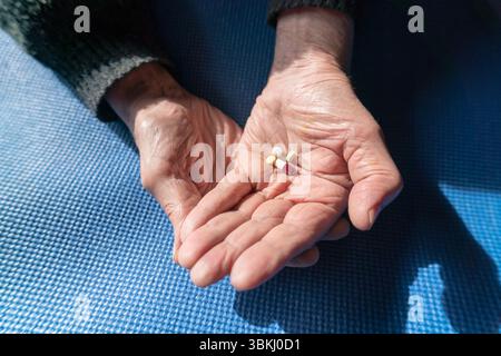Primo piano di mani anziane che tengono pillole medicinali su un tappetino blu, illuminato dalla luce naturale del sole Foto Stock