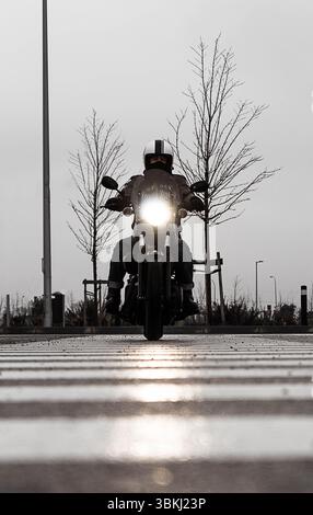 Un motociclista con un casco attraversa la città in una giornata di pioggia, con il suo faro che splende brillantemente su una strada bagnata. Foto Stock