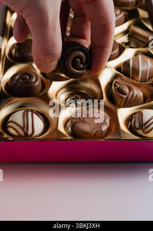 Una mano umana entra in una scatola d'oro di cioccolatini assortiti, raccogliendo una delle prelibatezze deliziose, con altri cioccolatini visibili Foto Stock
