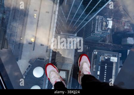 La foto mostra i piedi di una persona in piedi su un pavimento di vetro con una vista mozzafiato della città sottostante. I piedi della persona sono rivestiti di sneakers bianche Foto Stock
