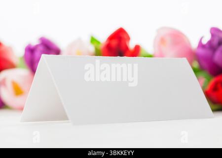 La scheda vuota in stile tenda dal design minimalista è posizionata su una superficie bianca con sfondo in tulipano sfocato Foto Stock