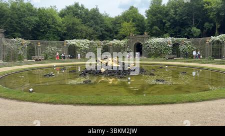Vista giardino con laghetto centrale con una scultura dorata, incorniciata da trellises floreali e lussureggianti alberi verdi. Foto Stock