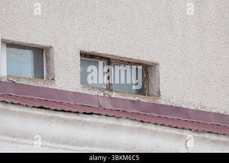 Una vista ravvicinata della parete esterna di un edificio in decadenza presenta due finestre, una intatta, una rotta sopra una linea rossa del tetto in metallo ondulato, che espone S. Foto Stock