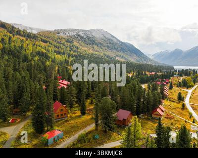 Una vista aerea raffigura un rifugio panoramico di montagna con capanne dal tetto rosso circondate da fitti boschi di pini e colori autunnali, con un lago nel dis Foto Stock