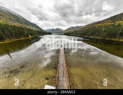 Un lungo molo di legno si estende nel lago Rakhmanovskoe, riflettendo cieli nuvolosi e montagne autunnali nelle sue acque calme Foto Stock
