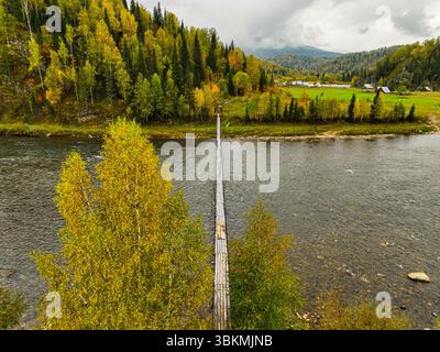 Un rustico ponte sospeso in legno attraversa un fiume, che conduce verso una vibrante foresta autunnale e colline lontane sotto un cielo nuvoloso Foto Stock