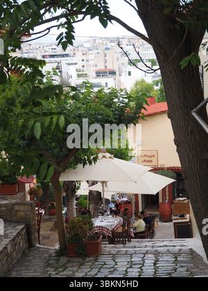 Immagine editoriale di un vivace ristorante all'aperto sotto ombrelloni bianchi a Plaka, Atene, Grecia, con persone che cenano in un ambiente ombreggiato dagli alberi. Foto Stock