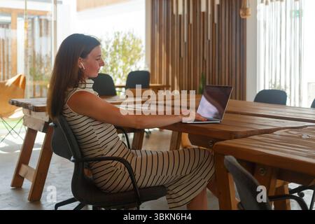 Una donna seduta su un tavolo di legno utilizza il proprio laptop in uno spazio di lavoro luminoso e contemporaneo pieno di luce naturale e vegetazione. Foto Stock