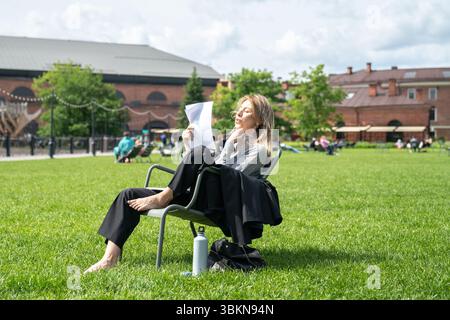 Donna scomoda che si sventola con fogli di carta come un ventilatore mentre si siede sulla sedia all'esterno sul prato. Foto Stock