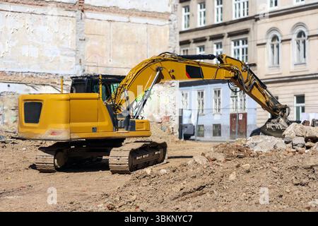 Un grande escavatore giallo si trova su un cantiere con mucchi di sabbia e macerie, con vecchi edifici visibili sullo sfondo. Foto Stock