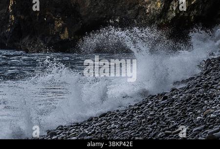 Una potente onda oceanica si schianta contro una costa rocciosa, creando una scena dinamica di acqua e pietra. La schiuma bianca dell'onda e gli spruzzi contrastano con il TH Foto Stock
