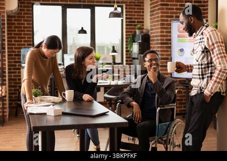 L'uomo afroamericano in sedia a rotelle gode di una conversazione spensierata con i suoi diversi colleghi durante una pausa. I colleghi multietnici ridono insieme in un ufficio a muro, condividendo storie. Foto Stock