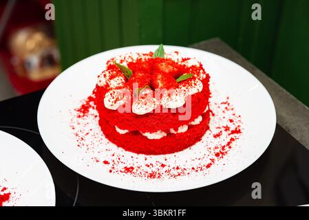 Mini torta di velluto rosso brillante ricoperta di fragole e panna, servita su un piatto bianco con sfondo pulito. Foto Stock