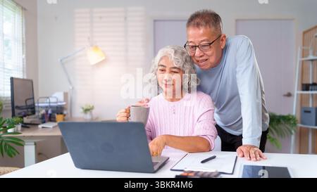 Una coppia asiatica più anziana si sta godendo la vita in pensione a casa, usando un notebook insieme e prendendo un caffè a casa mentre guarda il co Foto Stock
