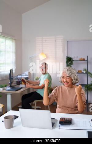 Una donna asiatica più anziana celebra allegramente il successo nel suo ufficio domestico con un notebook, mostrando la gioia dell'equilibrio della vita pensionistica per una coppia anziana. Foto Stock