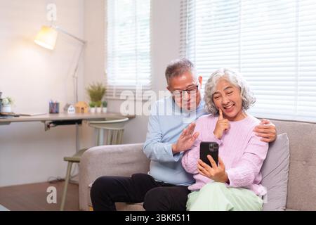 Felice coppia asiatica anziana che ha una piacevole videochiamata su uno smartphone a casa. Stanno godendo di una conversazione spensierata e sorridono insieme. Foto Stock