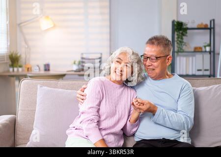Una coppia asiatica più anziana gode di un momento di pace sul proprio divano esprimendo profondo amore e cura. Questa immagine raffigura la bellezza dell'invecchiamento insieme con grazia Foto Stock