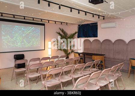 Moderno spazio eventi con schermo proiettore, file di sedie, illuminazione soffusa ed elementi decorativi per presentazioni o riunioni. Foto Stock