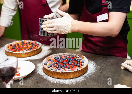 La persona che indossa i guanti cosparge di zucchero a velo su una crostata di fragole e mirtilli appena preparata durante la preparazione del dessert in cucina. Foto Stock