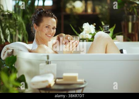 Una donna sorridente di mezza età gode di un lussuoso bagno di bolle, che tiene il tè. Capelli bagnati tirati indietro, si rilassa tra lussureggianti piante verdi e luci calde, e. Foto Stock