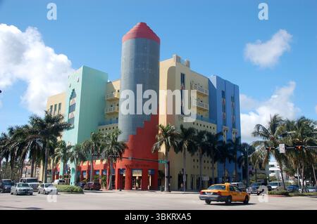 Edificio colorato in stile Art Deco con una torre a forma di matita su un angolo di strada soleggiata, circondato da palme e traffico a Miami Foto Stock