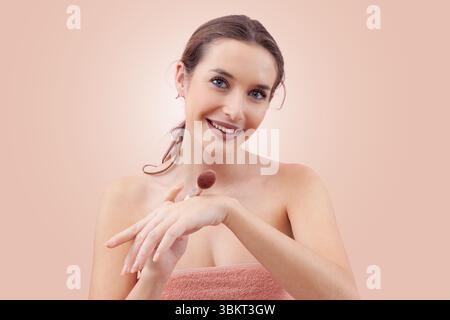Una giovane donna sorridente che tocca un pennello per il trucco sul dorso della mano prima di applicare il blush mentre guarda la fotocamera. Routine di bellezza pulita e minimalista Foto Stock
