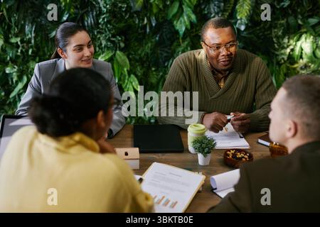 Gruppo eterogeneo di giovani adulti e professionisti di mezza età che discutono della strategia aziendale attorno a un tavolo, team multietnico che collabora con documenti, grafici e tablet digitali visibili Foto Stock