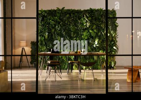 Area di lavoro vuota e moderna con grande scrivania in legno con laptop aperto e tazza da caffè, circondata da sedie a filo e lussureggiante parete verde illuminata da luce naturale Foto Stock
