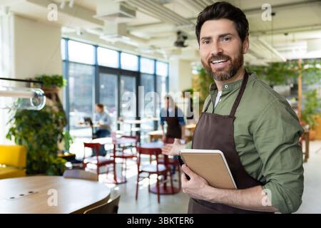Giovane barbuto in grembiule che guarda fiducioso mentre aspetta il cliente in pizzeria Foto Stock
