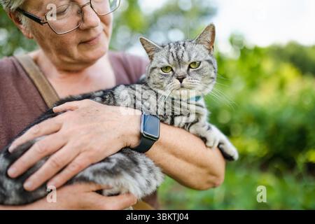 Una donna anziana tiene delicatamente un gatto Shorthair britannico in un giardino lussureggiante, entrambi guardando attentamente in lontananza in una giornata calda. Foto Stock
