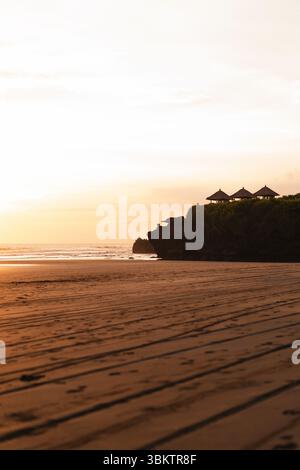 Tramonto dorato su una spiaggia tranquilla con tetti balinesi tradizionali Foto Stock