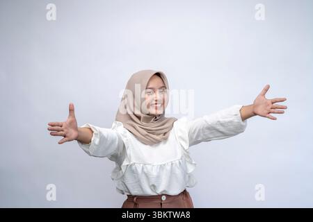 Mi dia Hug. Ritratto della giovane donna indonesiana positiva che indossa hijab che si allunga le mani a camera, felice sorridente Millennial apertura braccia per Em Foto Stock