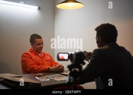 Giovane uomo caucasico adulto in tuta arancione della prigione seduta al tavolo degli interrogatori ammanettato, uomo nero di mezza età in tuta che lo intervista, video videocamera registrazione conversazione Foto Stock