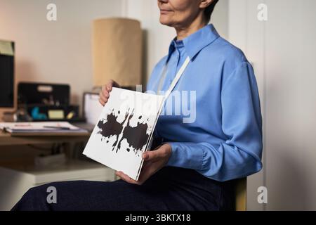 Donna caucasica di mezza età seduta in casa con la carta di prova del inkblot in mano, faccia parzialmente visibile, impegnata in una valutazione psicologica o sessione di terapia, sfondo dell'ufficio visibile Foto Stock